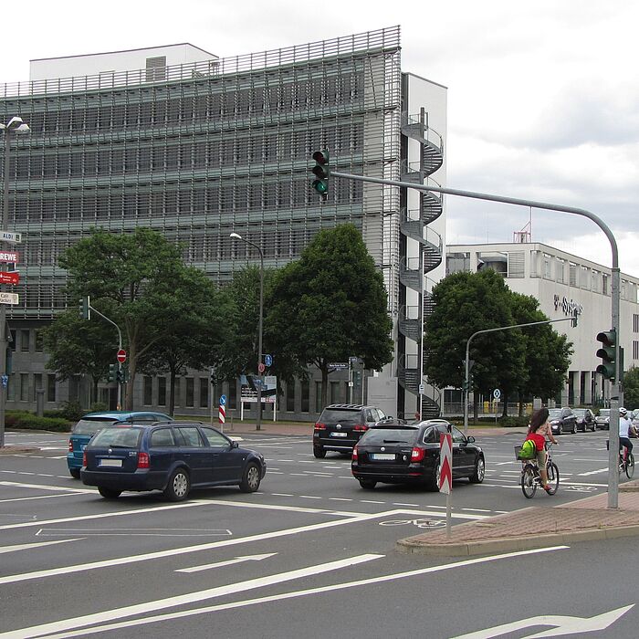 Kreuzung mit Abbiegestreifen und Lichtsignalanlage, zahlreiche Fahrzeuge und Fahrradfahrer, im Hintergrund hohe Gebäude