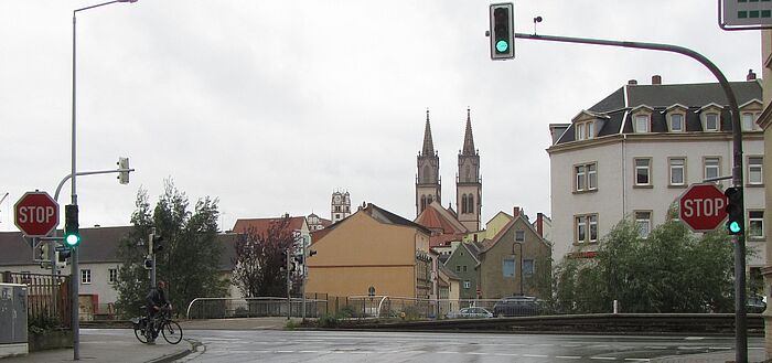 Straßeneinmündung mit Ampel und STOPP-Beschilderung, Radfahrer wartet, im Hintergrund Bebaung und Kirchtürme