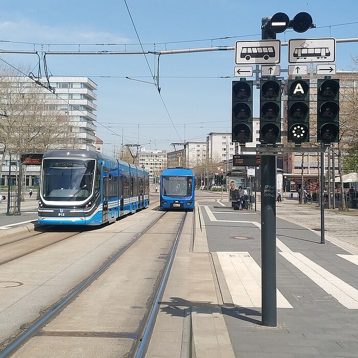 Straßenbahnhaltestelle mit zwei in gegensätzliche Richtung fahrenden Straßenbahnen, digitale Abfahrtstafel, LSA für Straßenbahn im Vordergrund