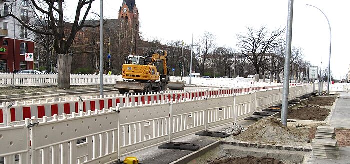 Baustelle mit Baustellenabsperrungen, Bagger, Gehweg mit offenen Erdflächen, Bebauung, Kirche im Hintergrund