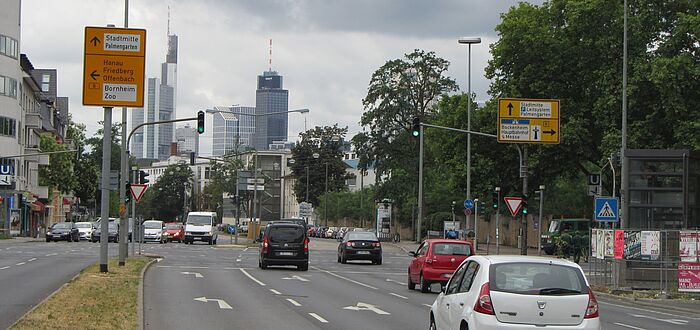mehrspurrige Kreuzung mit Abbiegestreifen und Lichtsignalanlage, zahlreiche Fahrzeuge, im Hintergrund Skyline Frankfurt am Main
