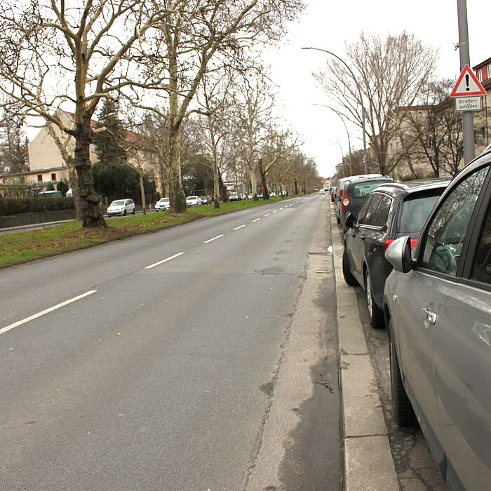 Bestandsituation im Bereich Fahrbahn und Mittelstreifen 2 streifige Fahrbahn mit Längsparkern und Bebauung im Hintergrund