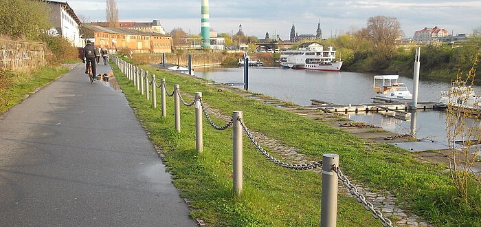 links Fahrradweg mit Radfahrer, Absperrung zum Neustädter Hafen, Hafen mit Schiff im Hintergrund