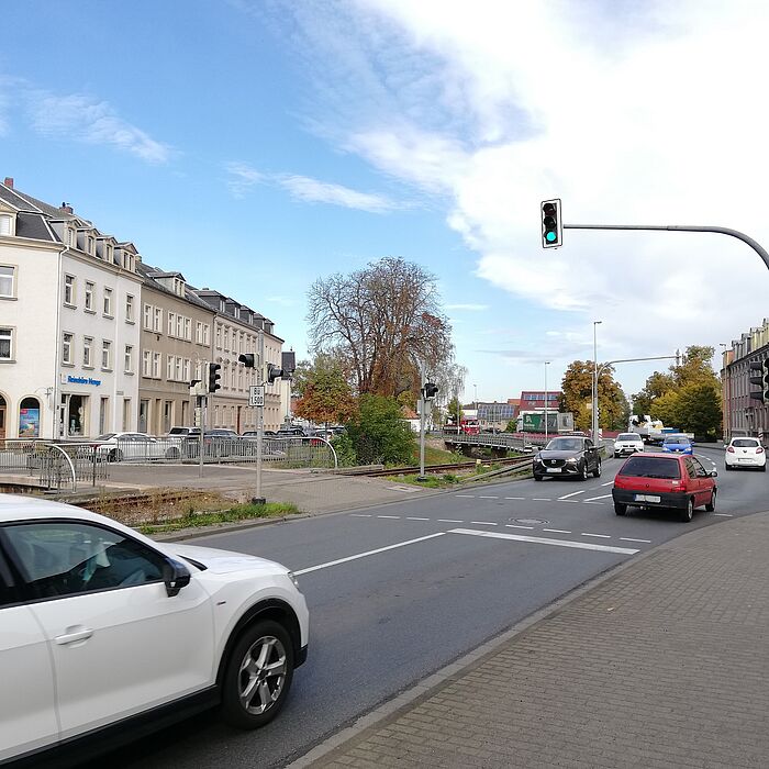 befahrene Straße mit Ampelkreuzung, links parallel verlaufenden Gleisen und Bahnübergang, rechts Bebauung